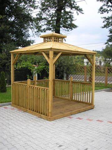 Drewniana altana ogrodowa z krzyżowymi zdobieniami, widoczna z zewnątrz, na tle zieleni i kostki brukowej.